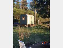 Kleines, hellgraues Gartenhäuschen mit Runddach und zwei Fenstern auf einer Holzplattform in einem grünen Garten, umgeben von Bäumen und Sträuchern. Im Vordergrund stehen eine weiße Liege auf einer gepflasterten Fläche und eine Feuerschale mit brennendem Feuer.