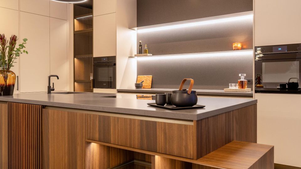 Moderne Wohnküche mit großer Kücheninsel aus Holz und grauer Arbeitsplatte, integriertem Spülbecken und Sitzbank. Helle, grifflose Schränke, eingebaute Backöfen, offene Regale mit indirekter LED-Beleuchtung und runde Deckenleuchte prägen den Raum.