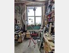 Eine Werkstatt mit einem großen Fenster, durch das Tageslicht einfällt. Links und rechts befinden sich Regale und Arbeitsflächen, die mit Werkzeugen, Materialien und Geräten gefüllt sind. Der Raum wirkt funktional und ist mit diversen Gegenständen ausgestattet, die auf handwerkliche Arbeiten hinweisen.