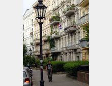 Straßenansicht mit Altbauten im Gründerzeitstil, verziert mit Balkonen voller Blumen. Eine gepflasterte Gehwegfläche mit Hecken, einer Gaslaterne in der Mitte und geparkten Autos links; im Hintergrund sind Personen zu sehen.