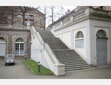 Eine breite Steintreppe mit weißen Balustraden führt zu einem höher gelegenen Pavillon mit großen Rundbogenfenstern. Links ist eine mit Efeu bewachsene Wand, und im Hintergrund sind Wohngebäude und kahle Bäume sichtbar.