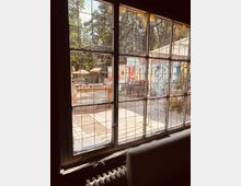 Blick aus einem großen, gitterverstärkten Fenster auf einen offenen Außenbereich mit bemalten Wänden, Holztischen, Stühlen und Sonnenschirmen; im Hintergrund sind Bäume sichtbar. Das Fenster gehört zu einem älteren Gebäude mit sichtbarem Heizkörper im Vordergrund.