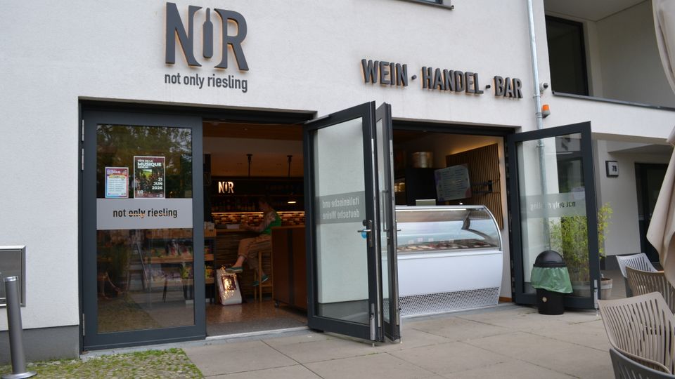 Eingangsbereich eines Weinhandels mit Bar („NUR – not only riesling“) in einem modernen, hell verputzten Gebäude, mit großen Glastüren und aufgesetzter Beschriftung „WEIN · HANDEL · BAR“. Durch die offenen Türen sind Theke und Kühlvitrine im Innenraum sichtbar; davor liegt eine gepflasterte Terrasse mit Sitzmöbeln.