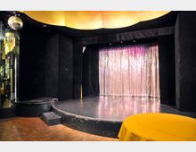 Kleiner Veranstaltungsraum mit einer Bühne, die von einem schwarzen Vorhangrahmen umgeben ist. Auf der Bühne befindet sich ein schimmernder, silberner Hintergrundvorhang; im Vordergrund sind ein Holzboden, ein runder Tisch mit gelbem Tuch und eine Diskokugel an der Decke sichtbar.