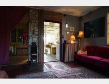 Gemütlicher Innenraum eines mit Holz und Stein gestalteten Wohnzimmers, ausgestattet mit einem roten Sofa, mehreren Lampen, Teppichen und dekorativen Kunstgegenständen. Im Hintergrund ist eine offene Tür, die in eine helle Küche mit gelbem Tisch führt.