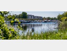 Ein ruhiger Hafen mit festgemachten Motorbooten und Yachten, umgeben von modernen Wohngebäuden und üppigem Grün im Vordergrund. Ein Schild mit „Privat“ steht im Wasser, und die Szene wird von Bäumen und Sträuchern eingerahmt.