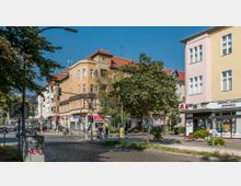 Straßenszene in einer Stadt mit mehrstöckigen Wohn- und Geschäftshäusern, darunter eine Apotheke im Erdgeschoss. Im Vordergrund befinden sich ein Baum, eine Fußgängerampel, ein Kreisverkehr sowie Gehwege mit Passanten und Radfahrern.