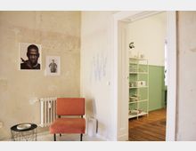 Ein schlichter Wohnraum mit einer abgenutzten, beigefarbenen Wand, an der zwei gerahmte Schwarz-Weiß-Fotografien hängen. Ein orangefarbener Stuhl steht vor einem weißen Heizkörper, daneben ein kleiner, runder Beistelltisch. Durch eine weiße Türöffnung ist ein Raum mit grün gestrichenen Wänden, Holzboden und einem weißen Regal mit Geschirr sichtbar.