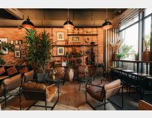 Gemütliches Café mit industriellem Design, Ziegelwänden, hängenden Lampen und modernen Ledersesseln. Dekorative Regale mit Pflanzen, Bildern und Vintage-Objekten ergänzen die Einrichtung, während große Fenster Tageslicht hereinlassen.