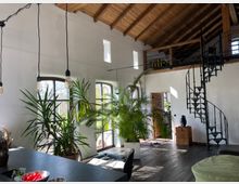 Wohnraum mit hohen, weißen Wänden und sichtbarem Holzbalkendach. Der Raum ist mit großen, bogenförmigen Fenstern, Pflanzen, einer schwarzen Wendeltreppe aus Metall und Holzmöbeln ausgestattet; Tageslicht fällt durch die Fenster.