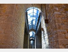 Steinerner Gang in einem historischen Gebäude mit massiven, rauen Natursteinwänden und einem schmalen, lichtdurchlässigen Glasdach, das von Metallstreben gestützt wird. Die Struktur zeigt bogenförmige Durchgänge und Lichteinfall von oben.