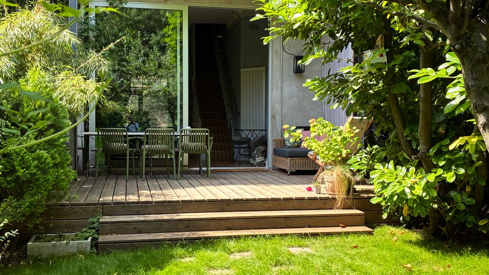 Holzterrasse im Garten vor einer modernen Hausfassade mit großer Glasschiebetür und Blick in ein Treppenhaus. Auf der Terrasse stehen ein Esstisch mit Stühlen sowie eine Sitzbank, umgeben von dichtem Grün, Sträuchern und einem großen Baum.