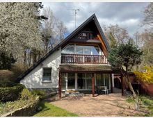Zweistöckiges Einfamilienhaus mit Satteldach und holzverkleideter Dachgaube. Das Gebäude ist von einem Garten mit blühenden Bäumen, Sträuchern und einer gepflasterten Terrasse umgeben; im Hintergrund befindet sich ein Wald.