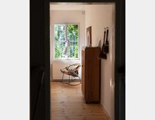 Blick in ein helles Zimmer mit Holzboden, einem großen Fenster mit Blick ins Grüne, einem Rattansessel mit buntem Kissen und einem schmalen Holzschrank. An der Wand hängen Pinsel und ein gerahmtes Bild lehnt am Schrank.