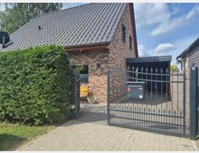Einfamilienhaus mit Ziegelfassade und steilem, grauem Ziegeldach, davor ein schmiedeeisernes Tor mit Briefkasten und Zufahrt zu einem überdachten Carport. Links steht eine hohe, dicht geschnittene Hecke, und der Gehweg führt zum Eingang unter dem Dachüberstand.