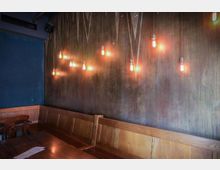 Holzvertäfelte Sitzecke mit einer braunen Wand, an der mehrere freihängende Glühbirnen in warmem Licht leuchten, und eine angrenzende dunkelblaue Wand. Im Vordergrund ein Holztisch mit darauf liegenden Papieren.