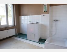 Unrenovierter Sanitärraum mit weiß gefliesten Wänden, einem gefliesten Waschbeckenblock und einer Duschwanne mit Handbrause. Links befindet sich ein großes Fenster über einem Heizkörper; der Boden ist teils gefliest und teils roh, mit sichtbaren Installationsspuren an der Wand.