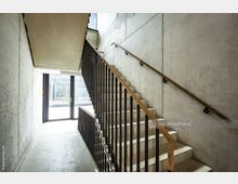 Betontreppenhaus mit modernem Design, bestehend aus einer geraden Treppe mit Holzhandläufen und schwarzen Metallstreben. Im Hintergrund befindet sich eine Glastür mit Blick nach draußen und viel einfallendem Tageslicht.