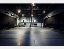 Ein großer, leerer Probenraum mit dunklen Wänden und schwarzem Bodenbelag, beleuchtet von mehreren Deckenstrahlern. Im Hintergrund befinden sich eine Metalltreppe, Arbeitsflächen, technisches Equipment und weiße Türen.