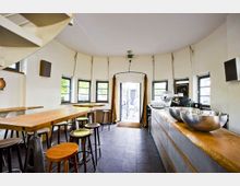 Moderner Innenraum eines Cafés oder einer Bar mit runden Wänden, mehreren Fenstern und weiß gestrichenen Wänden. Zu sehen sind ein langer Holztresen mit großen Metallschüsseln, hohe Tische mit Hockern sowie eine Wendeltreppe im linken Bildrand. Im Hintergrund führt eine Tür nach draußen.