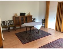 Esszimmer mit Holzmöbeln, darunter ein geschwungener Tisch mit weißer Tischdecke, vier gepolsterte Stühle im antiken Stil und eine verzierte Kommode mit Glasfront. Der Raum verfügt über Laminatboden, Teppiche mit orientalischem Muster und eine weiße Wand mit Holzelementen.