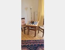 Ein kleiner Holztisch mit einem Schachbrett und Figuren steht zwischen zwei Holzstühlen, in einem Raum mit hellem Holzboden. Im Hintergrund sind ein gelber Vorhang, ein Heizkörper und eine Stehlampe zu sehen.