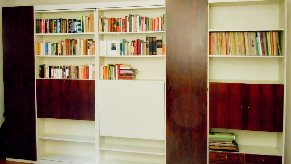 Große, wandfüllende Einbauregale in Weiß mit dunkelbraunen Holzschiebetüren, teils geöffnet und mit Büchern sowie Aktenordnern gefüllt. Unten befinden sich geschlossene Schrankfächer und Schubladen, oben mehrere offene Regalböden.