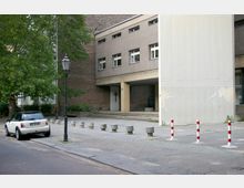 Ansicht eines modernen Gebäudes mit einfacher, rechteckiger Fassade und mehreren kleinen Fenstern, das an eine gepflasterte Fläche mit Betonpollern und rot-weiß markierten Absperrungen grenzt. Links im Bild steht ein geparktes Auto neben Bäumen und einer Straßenlaterne, während rechts eine kleine Treppe zum Eingang des Gebäudes führt.