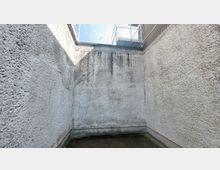 Kleiner, betonierter Innenhof mit hohen, rau verputzten Wänden. Oben ist ein Drahtgitter und ein Teil eines benachbarten Gebäudes zu sehen.