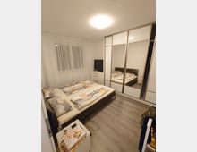 Ein modern eingerichtetes Schlafzimmer mit einem Doppelbett, dessen Bettwäsche ein Cartoon-Muster aufweist. Im Raum befinden sich ein weißer Kleiderschrank mit Spiegeltüren, eine Kommode mit einem Fernseher sowie ein kleiner Nachttisch mit Büchern. Der Boden ist mit hellem Holzlaminat gestaltet, und das Fenster ist mit weißen Gardinen bedeckt.