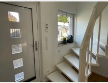 Weiß gestrichener Eingangsbereich mit moderner Haustür mit matten Glaseinsätzen, daneben ein Fenster mit Pflanzen- und Dekorationsobjekt auf der Fensterbank. Rechts davon führt eine schmale weiße Treppe nach oben.