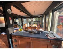 Offener Wohnraum mit einer modernen Küche, Holzbalken an der Decke und großen Fensterfronten, die viel Tageslicht hereinlassen. Im Hintergrund befinden sich ein Esstisch mit Stühlen und ein Blick auf eine Terrasse mit weiteren Sitzgelegenheiten.