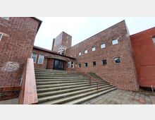 Backsteingebäude mit breiter Treppe, die zu einem Eingang führt, der von geschwungenen Wänden eingerahmt ist. Der Bau ist durch rote Klinkersteine, rechteckige Fenster und einen Turm im Hintergrund gekennzeichnet.
