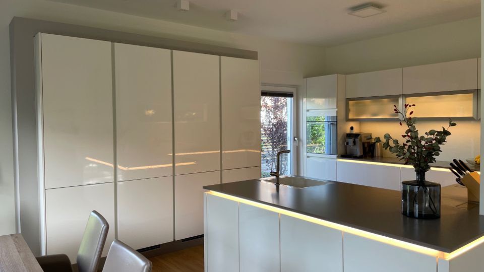 Moderne Küche mit weißen Hochglanzfronten, einer Kochinsel mit dunkler Arbeitsplatte und integriertem Spülbecken. Im Hintergrund befinden sich hohe Schränke, eine Kaffeemaschine, und eine Tür, die zu einem Gartenbereich führt; die Beleuchtung ist dezent in die Möbel integriert.