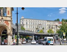 Straßenszene in einer Stadt mit historischem Gebäude auf der linken Seite, einer erhöhten Bahntrasse aus Metall in der Mitte und einem großen Wohnhaus im klassizistischen Stil im Hintergrund. Im Vordergrund sind Autos, ein Lieferroller und Fußgänger an einer Ampelkreuzung zu sehen.