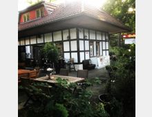 Fachwerkgebäude mit einem überdachten Außenbereich, in dem Tische und Stühle auf einer Terrasse stehen, umgeben von Pflanzen und Grün. Im Hintergrund befinden sich Fenster und ein Eingang, die zu einem Café oder Restaurant gehören könnten.