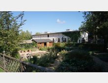 Großer Garten mit Blumenbeeten, einem Holzzaun und einem kleinen Holzschuppen im Vordergrund; im Hintergrund ein langes, weiß verputztes Gebäude mit bogenförmigen Fenstern und teilweise bewachsenen Wänden, umgeben von Bäumen unter klarem Himmel.