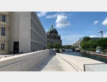 Fußweg entlang eines modernen Gebäudes aus hellem Stein mit rechteckigen Fenstern, links angrenzend ein historisches Gebäude. Im Hintergrund der Berliner Dom mit grüner Kuppel, rechts ein Fluss mit Uferpromenade und Bäumen.