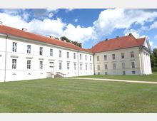 Helles, zweistöckiges Barockgebäude mit rotem Satteldach und weißen Fassaden, großen Fenstern und einem Eingangsbereich mit einer kleinen Treppe. Das Gebäude steht inmitten einer Rasenfläche unter einem blauen, leicht bewölkten Himmel.