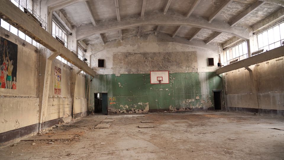 Verlassene Turnhalle mit hohem, gewölbtem Betondeckengebälk und großen Fensterbändern entlang der Seitenwände. An der Stirnwand hängt ein Basketballkorb vor abblätternder grüner Farbe, der Boden ist staubig und beschädigt, mit offenen Türdurchgängen an beiden Seiten.