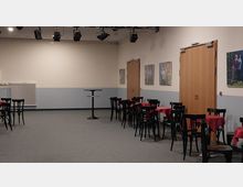 Ein schlichter Veranstaltungsraum mit Tischen und Stühlen, die mit roten Tischdecken dekoriert sind. An der hellen Wand hängen gerahmte Bilder, und es gibt zwei geschlossene Holztüren sowie an der Decke montierte Strahler.