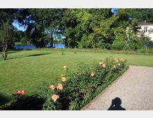 Ein gepflegter Garten mit einer großen Rasenfläche, einer Reihe blühender Rosen entlang eines Kieswegs und einem angrenzenden See im Hintergrund. Im hinteren Bereich sind dichte Bäume und ein Teil eines weißen Gebäudes sichtbar.
