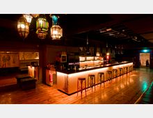 Eine stilvolle Bar mit langem Tresen, beleuchtet durch warmes, indirektes Licht, und mehreren Barhockern aus Holz. Die Decke ist mit bunten, orientalisch anmutenden Lampen geschmückt, während der Boden aus poliertem Holz besteht; im Hintergrund sind abstrakte Wandzeichnungen und ein kleiner Durchgang sichtbar.