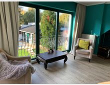 Wohnzimmer mit Holzboden, türkisfarbenen Wänden und großen Fenstern, die auf einen Garten mit Bäumen blicken. Im Raum stehen zwei gepolsterte Sessel, ein kleiner Holztisch und beige Vorhänge rahmen die Fenster ein.