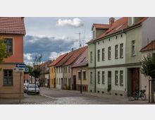 Eine ruhige Straße mit Reihen von zweistöckigen Wohnhäusern, die überwiegend mit pastellfarbenen Fassaden und roten Ziegeldächern versehen sind. Im Vordergrund stehen ein Straßenschild für eine Einbahnstraße, parkende Autos und ein Fahrrad.