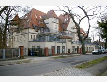 Mehrstöckiges Gebäude im historischen Stil mit hellen Fassaden und roten Satteldächern, teilweise mit Balkonen und großen Fenstern. Vor dem Gebäude verläuft eine asphaltierte Straße mit parkenden Autos, umgeben von Bäumen und Gehwegen.