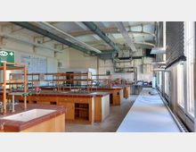 Ein Chemielabor mit mehreren hölzernen Labortischen, die mit Spülbecken, Wasserhähnen und Gasanschlüssen ausgestattet sind. An den Wänden befinden sich technische Geräte und Leitungen, während durch große Fenster auf der rechten Seite Tageslicht einfällt.