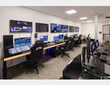 Moderner Kontrollraum mit mehreren Arbeitsplätzen, ausgestattet mit Monitoren, Schaltpulten und Computern. An der Wand hängen zahlreiche große Bildschirme, auf denen verschiedene Videoansichten zu sehen sind.