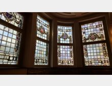 Große, farbige Buntglasfenster in einem geschwungenen Raum mit hohen Decken, verziert mit Wappen und dekorativen Mustern aus Ketten und floralen Elementen. Die Fenster sind geteilt in quadratische Segmente mit einer Mischung aus mattem und klarem Glas.