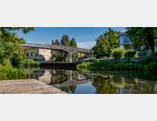 Eine Fußgängerbrücke mit moderner Metallkonstruktion überspannt einen kleinen Kanal, dessen ruhige Wasseroberfläche die umliegenden Bäume und Gebäude spiegelt. Im Hintergrund sind ein graffitibedecktes Gebäude und eine Grünfläche mit Bäumen sowie ein modernes Gebäude mit Glasfront zu sehen.